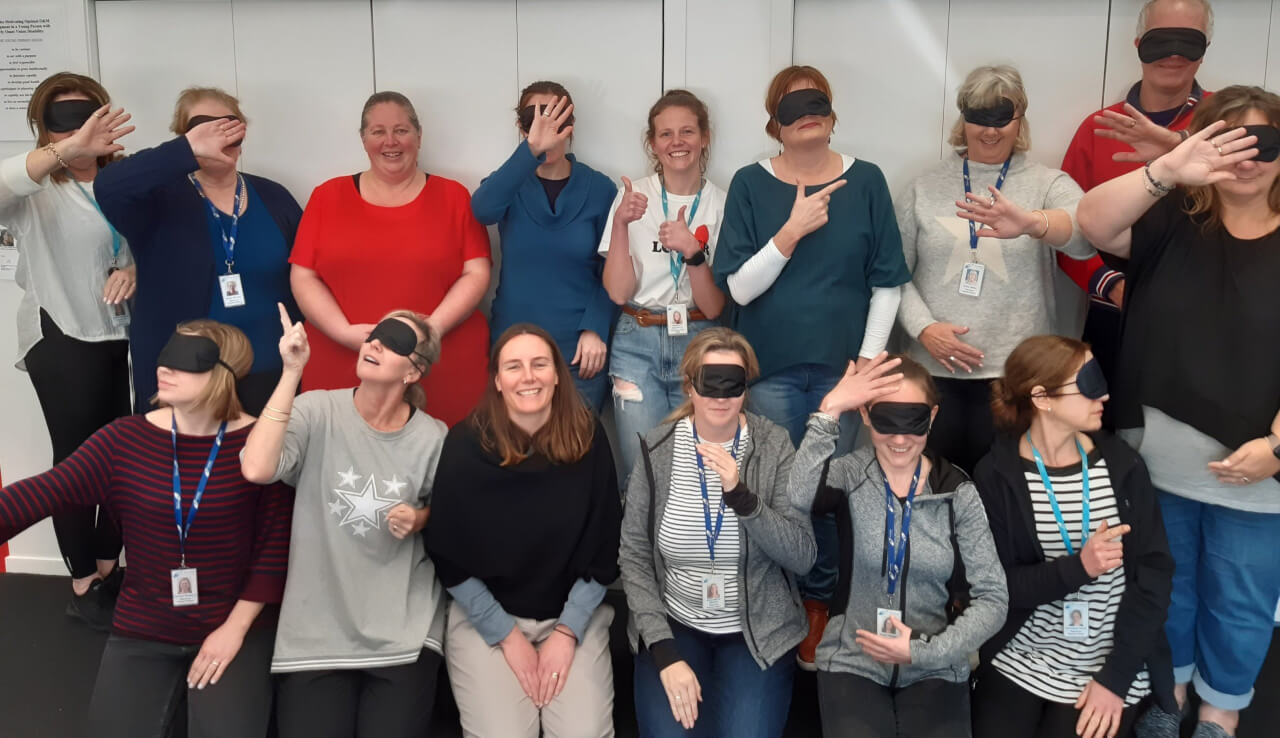 Figure 1 - Group photo of the Wellington VRC team, most wearing blindfolds, posing for the camera using guiding or trailing positions they have learned on the DOM Supporter course
