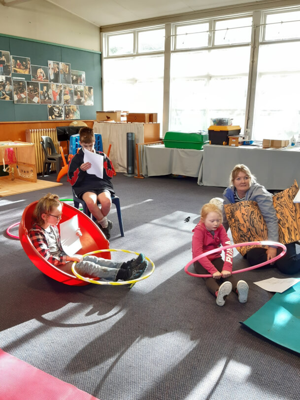 Figure 1 - A learner is sitting in a chair, another one is in a spinning toy, another one on the floor with a hula hoop with an adult reading and learning Japanese phrases