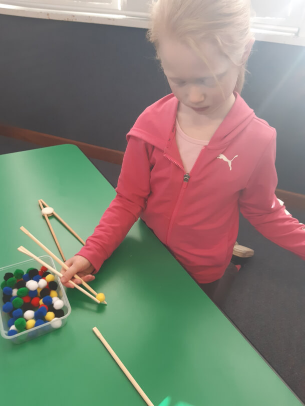 Figure 2 - Learner is sitting on the floor at a table using chopsticks to pick up fuzzy balls