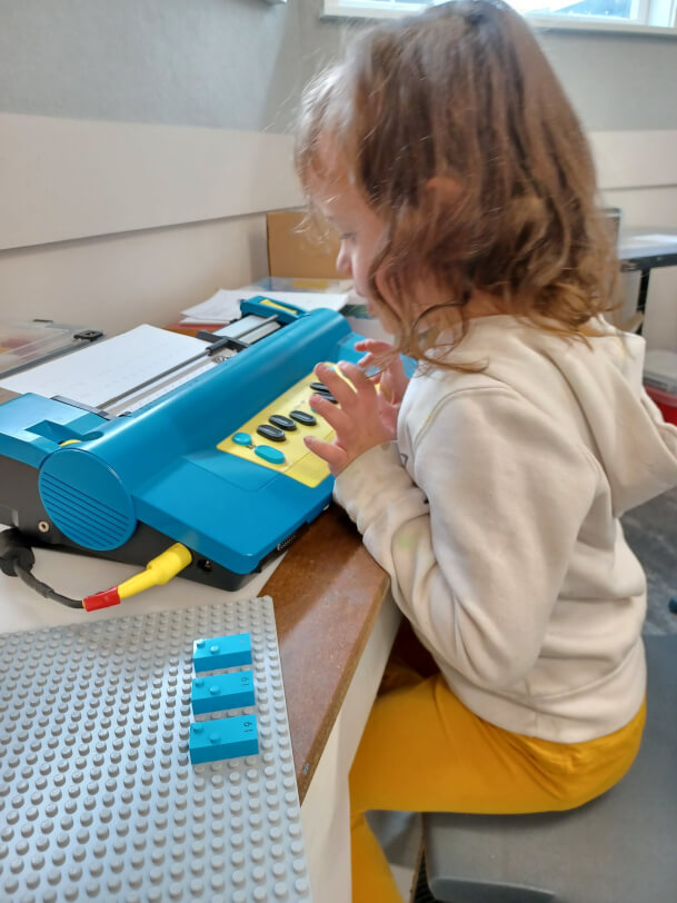 Figure 2 - Child is learning the letter ‘i’. Lego Braille Bricks are set up on a baseboard and she is brailling these on the Mountbatten.
