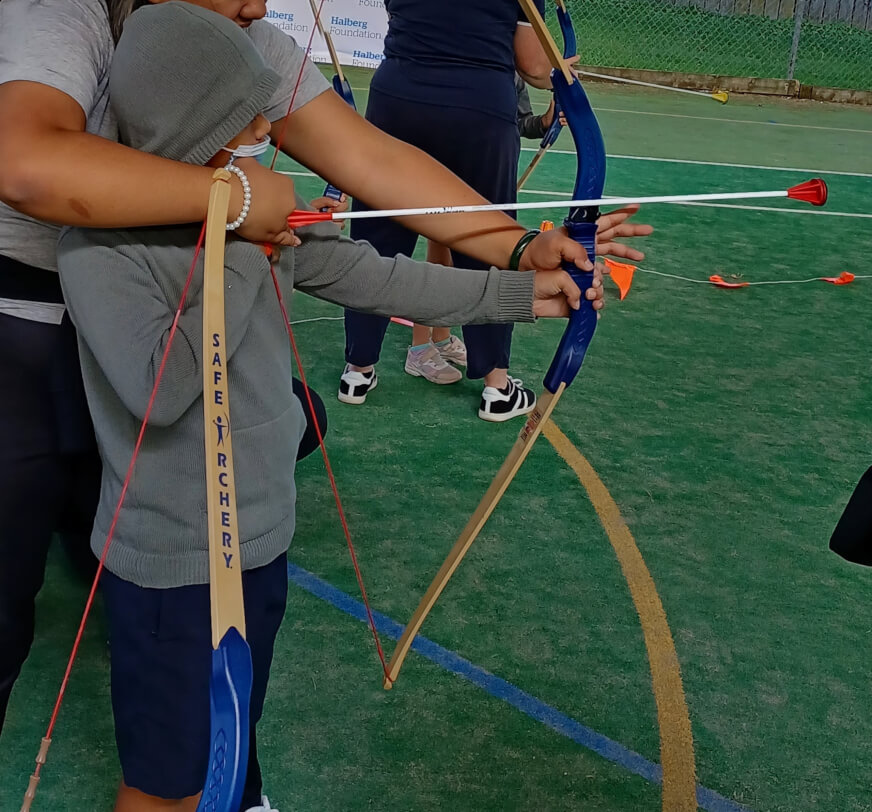 Figure 1 - Ākonga is standing with a bow and arrow in position learning how to shoot at the Halberg Sports Day. An adult is standing behind the ākonga helping hold the equipment