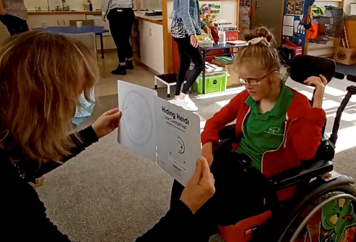 Figure 1 - Resource Teacher of Vision holding 2 square assessment cards in-front of 12 year old girl sitting in a wheelchair