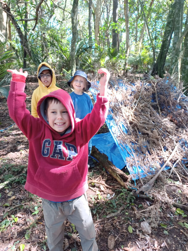 Figure 1 - Boy in red smiles with hands in the air, stood in front of his 2 smiling friends and a tarpaulin covered in sticks and leaves